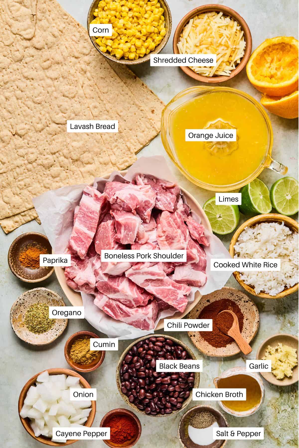 An overhead view of labeled ingredients for a Carnitas Burrito recipe, including boneless pork shoulder, lavash bread, corn, shredded cheese, orange juice, limes, white rice, black beans, onion, garlic, spices, chicken broth, and salt & pepper.