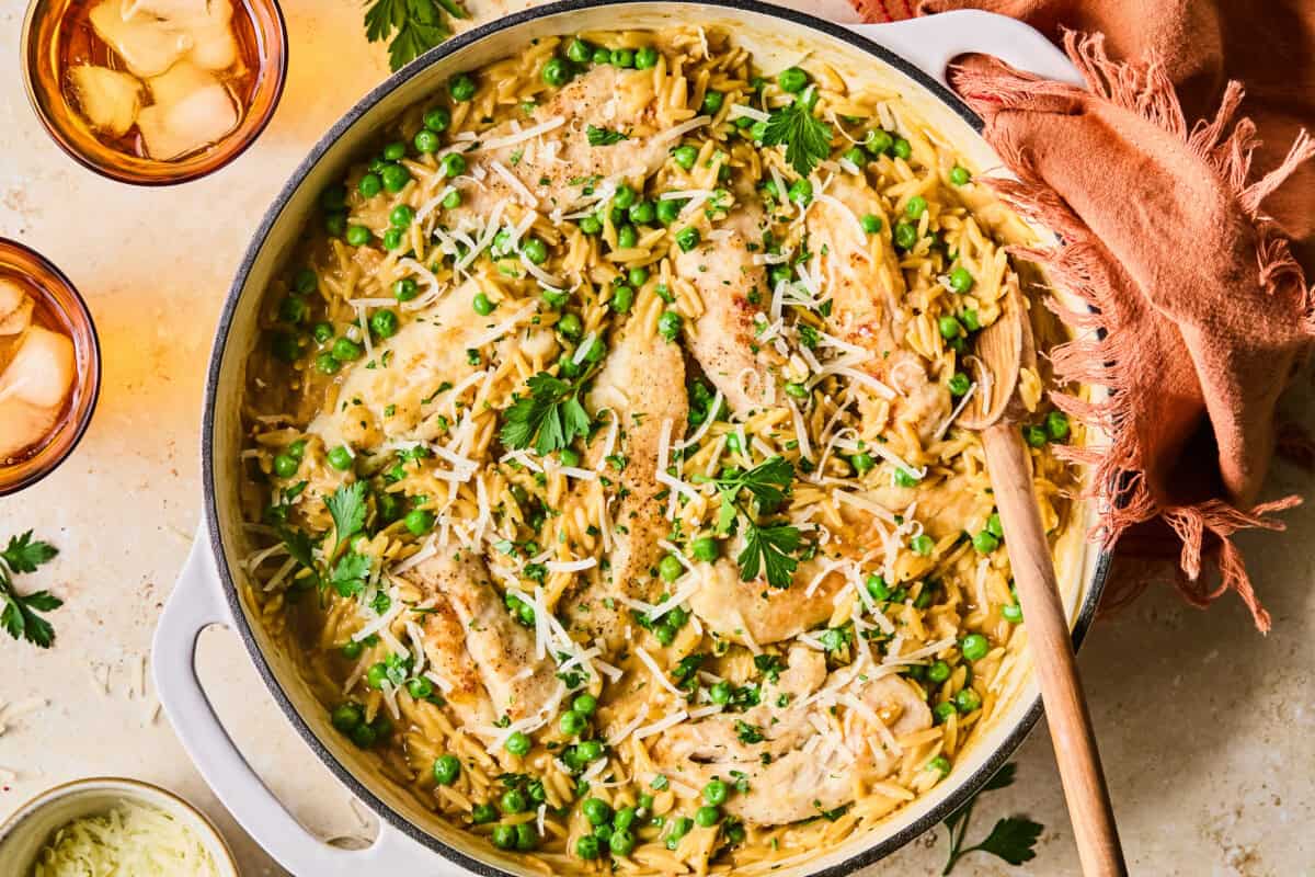 A large white pot filled with creamy chicken and orzo pasta, topped with chicken and peas, shredded cheese, and fresh parsley. A wooden spoon rests nearby with an orange napkin; iced drinks and herbs fill the background.
