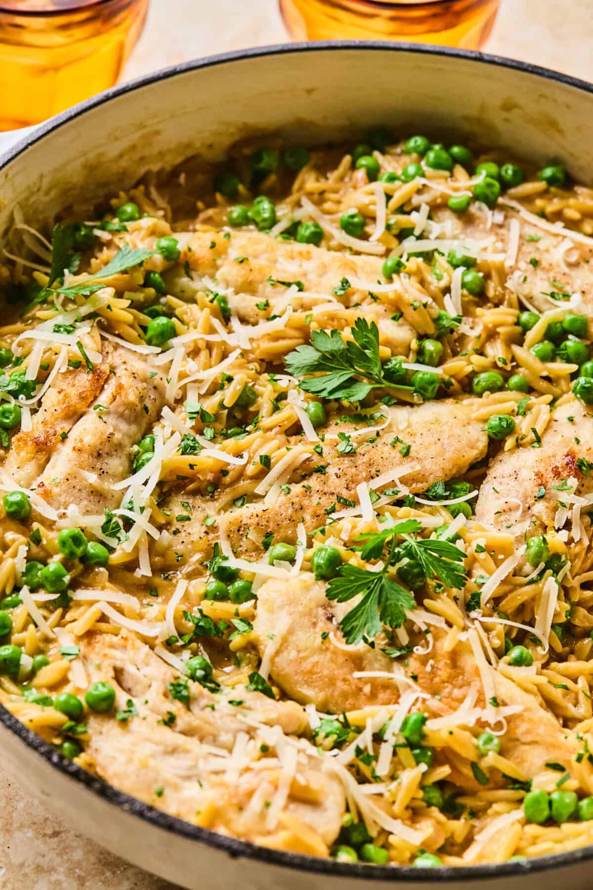 A skillet filled with cooked chicken and peas, orzo pasta, grated cheese, and fresh parsley, with two glasses of amber-colored drink in the background.