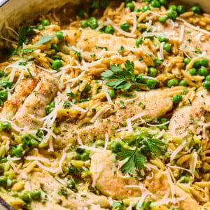 A skillet filled with cooked chicken and peas, orzo pasta, grated cheese, and fresh parsley, with two glasses of amber-colored drink in the background.