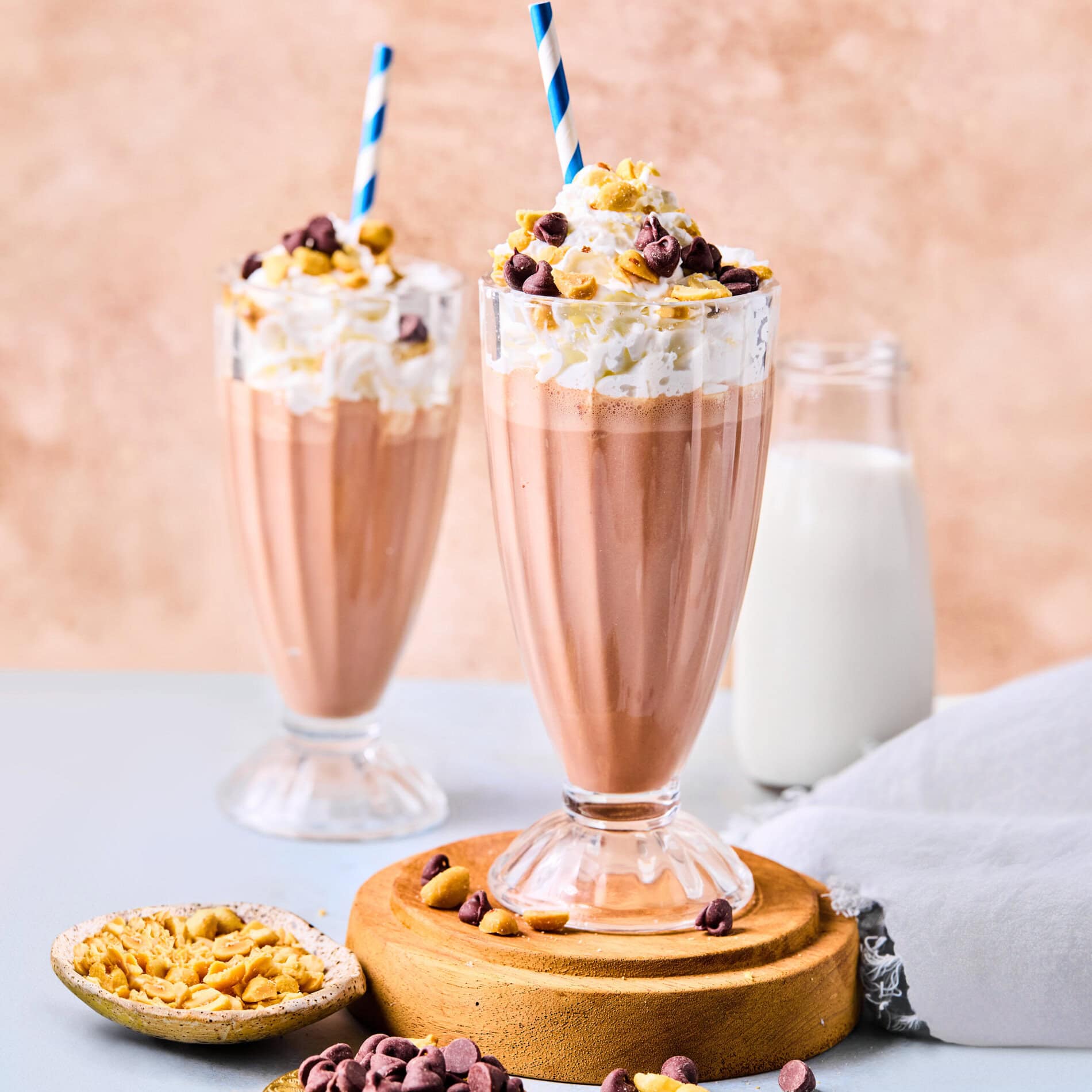 Two chocolate milkshakes in tall glasses topped with whipped cream, chopped nuts, and chocolate chips, each with a blue-striped straw—ideal for those seeking a smoothie recipe without banana. A bottle of milk and bowls of nuts and chips sit nearby.