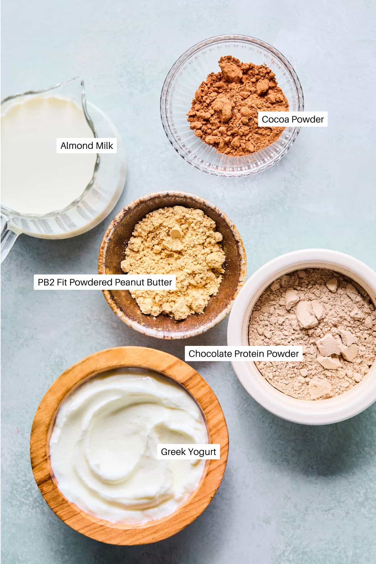 Five labeled ingredients—almond milk, cocoa powder, powdered peanut butter, chocolate protein powder, and Greek yogurt—are arranged in small bowls and a glass on a light surface, viewed from above for a smoothie recipe without banana.