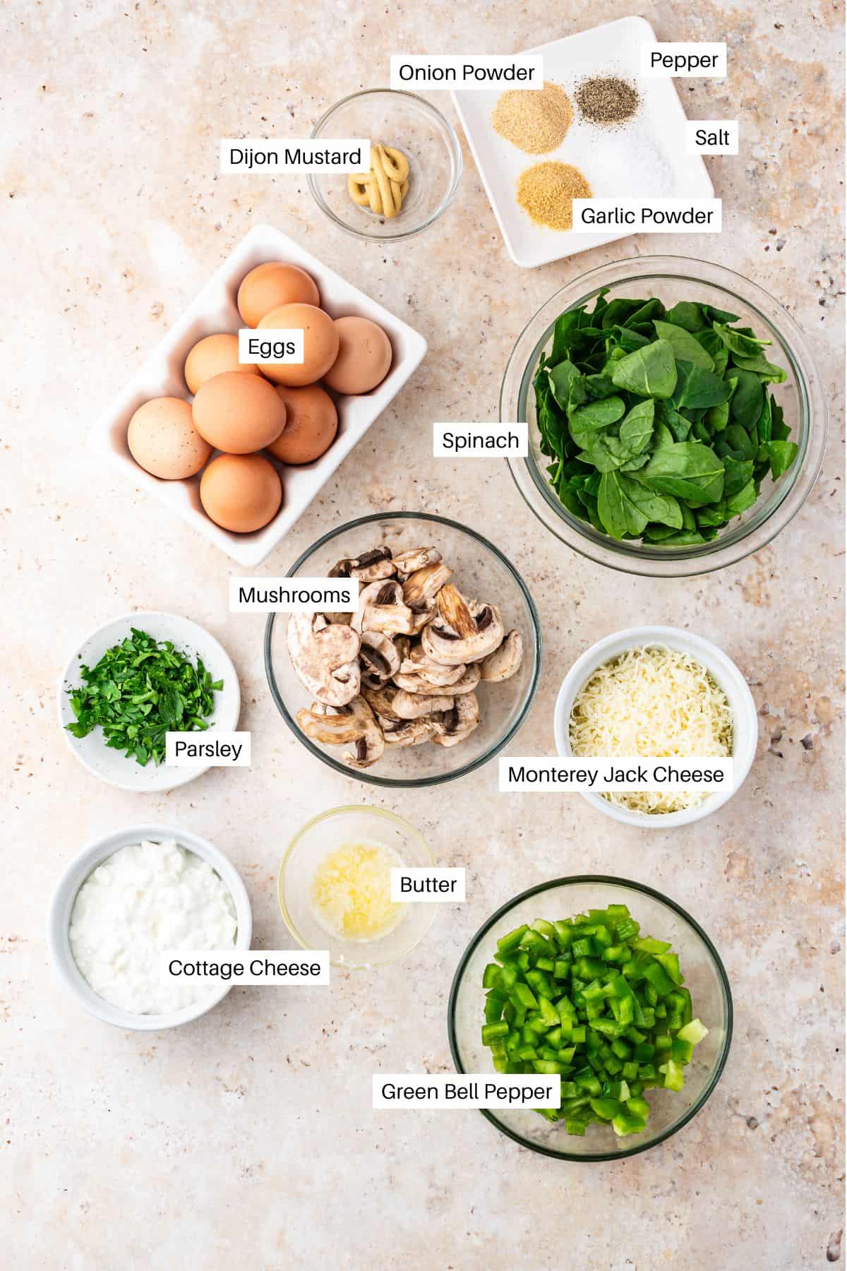 Overhead view of labeled ingredients for a cottage cheese egg casserole, featuring eggs, spinach, mushrooms, green bell pepper, Monterey Jack cheese, cottage cheese, parsley, butter, Dijon mustard, onion powder, garlic powder, salt, and pepper.