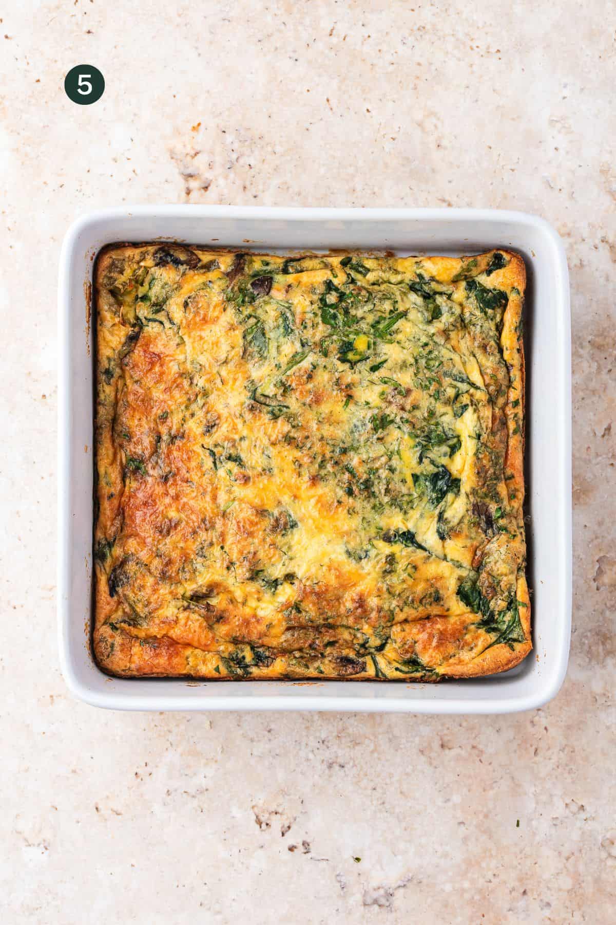 A baked cottage cheese egg casserole with spinach and cheese in a white square dish, resting on a light beige stone surface. A dark green circle with the number 5 appears in the upper left corner.