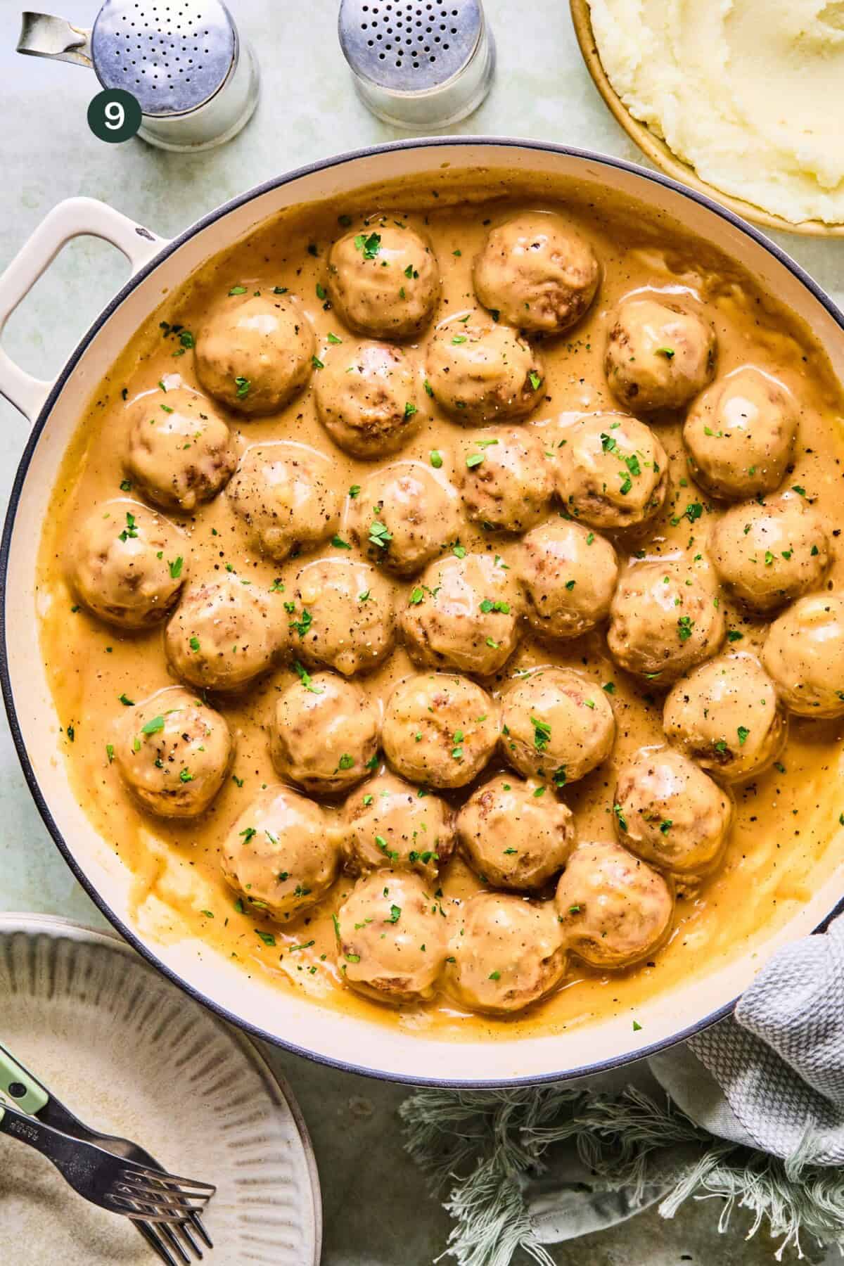 A white pan filled with meatballs and gravy, the meatballs covered in creamy brown sauce and garnished with chopped parsley. Nearby are plates, a fork, mashed potatoes, and salt and pepper shakers.