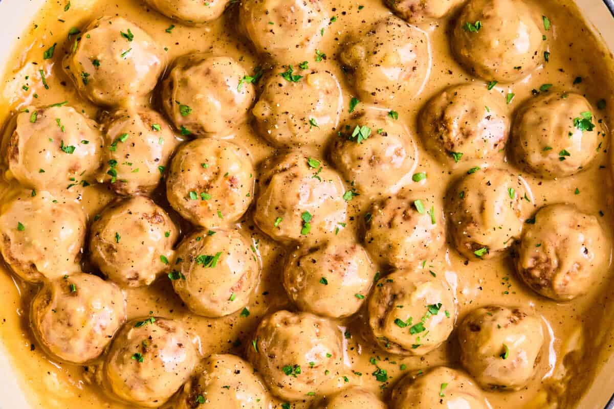 A close-up of meatballs and gravy, with the meatballs covered in creamy brown sauce and garnished with chopped parsley. They’re arranged closely together in a pan, making the dish look hearty and inviting.