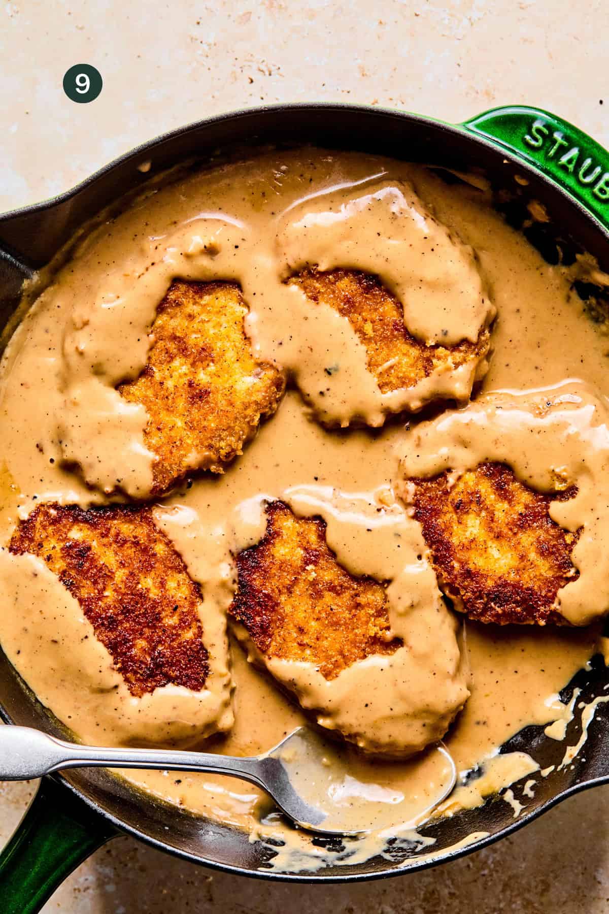 Five golden-brown breaded pork chops are partially covered in creamy gravy inside a black skillet, with a silver serving spoon resting at the edge.