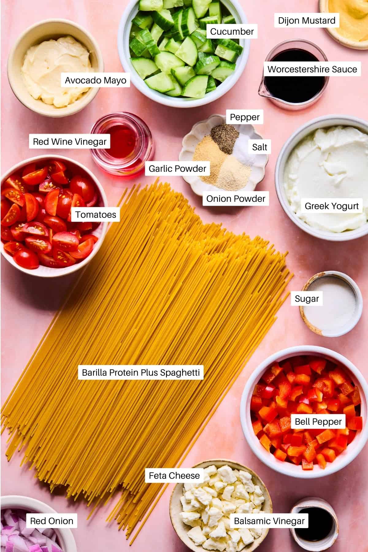 Ingredients for a spaghetti pasta salad are arranged on a pink surface, featuring uncooked spaghetti, chopped cucumber, tomatoes, red onion, bell pepper, feta cheese, Greek yogurt, avocado mayo, vinegars, and seasonings in bowls.