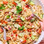 A large shallow bowl filled with spaghetti pasta salad, mixed with chopped cucumbers, cherry tomatoes, red onions, ham, herbs, and crumbled cheese. Two hands are serving the spaghetti pasta salad with wooden utensils.