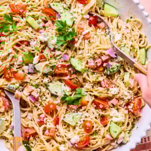 A large shallow bowl filled with spaghetti pasta salad, mixed with chopped cucumbers, cherry tomatoes, red onions, ham, herbs, and crumbled cheese. Two hands are serving the spaghetti pasta salad with wooden utensils.