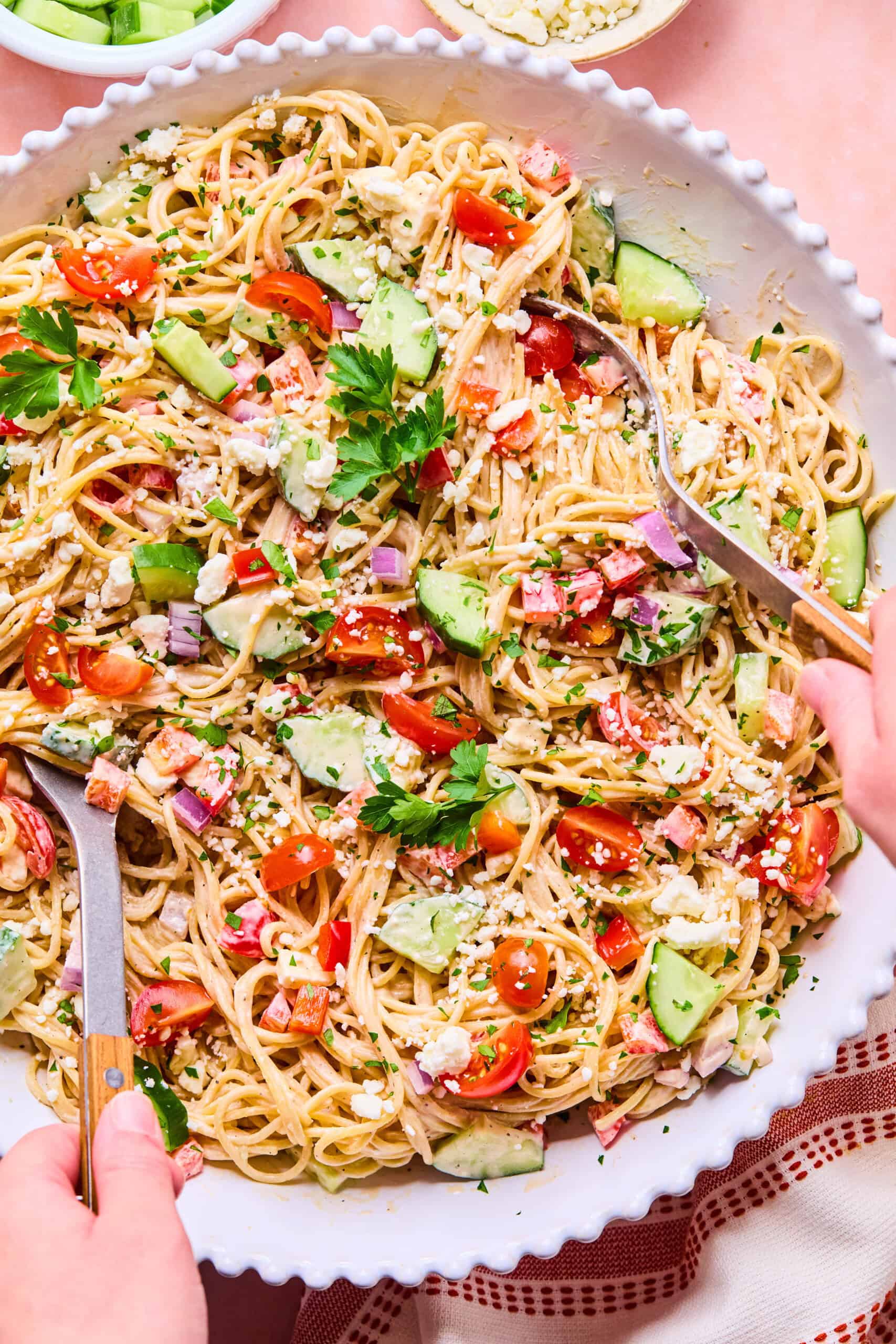 A large shallow bowl filled with spaghetti pasta salad, mixed with chopped cucumbers, cherry tomatoes, red onions, ham, herbs, and crumbled cheese. Two hands are serving the spaghetti pasta salad with wooden utensils.