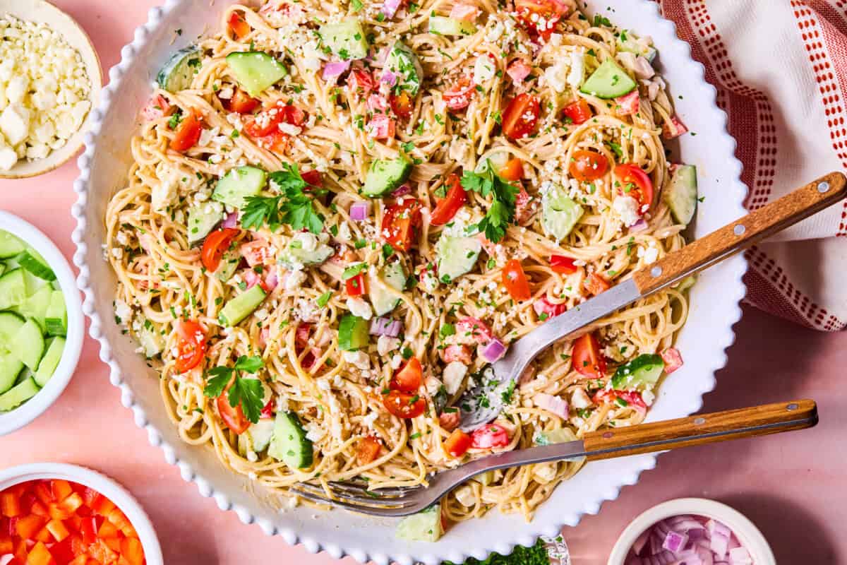 A large bowl of spaghetti pasta salad with cherry tomatoes, avocado, red onion, parsley, and crumbled cheese is served with wooden utensils. Small bowls of ingredients surround the dish on a pink surface.
