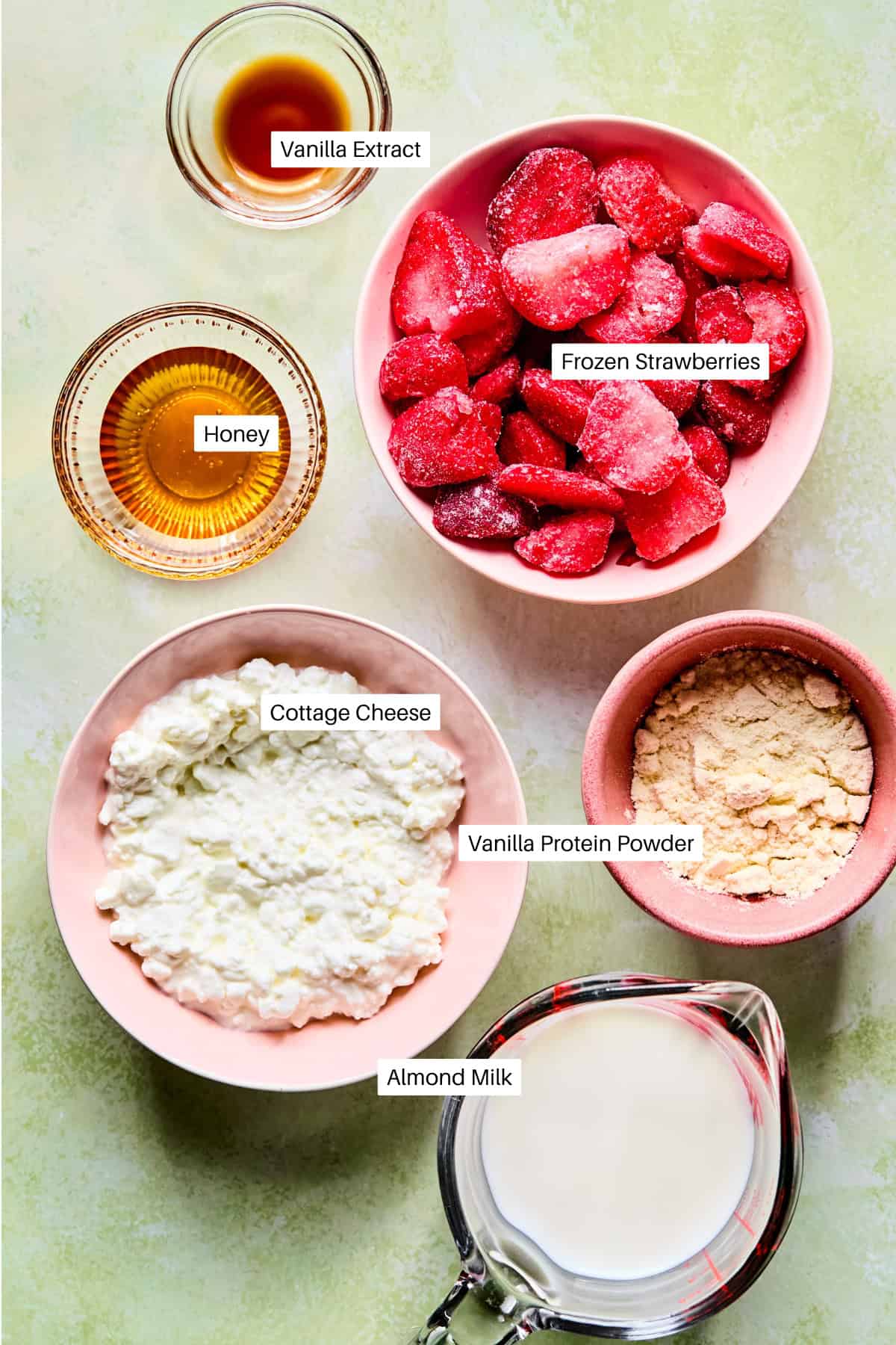 A flat lay of labeled ingredients for a strawberry protein shake: vanilla extract, honey, frozen strawberries, cottage cheese, vanilla protein powder, and almond milk on a light green background.