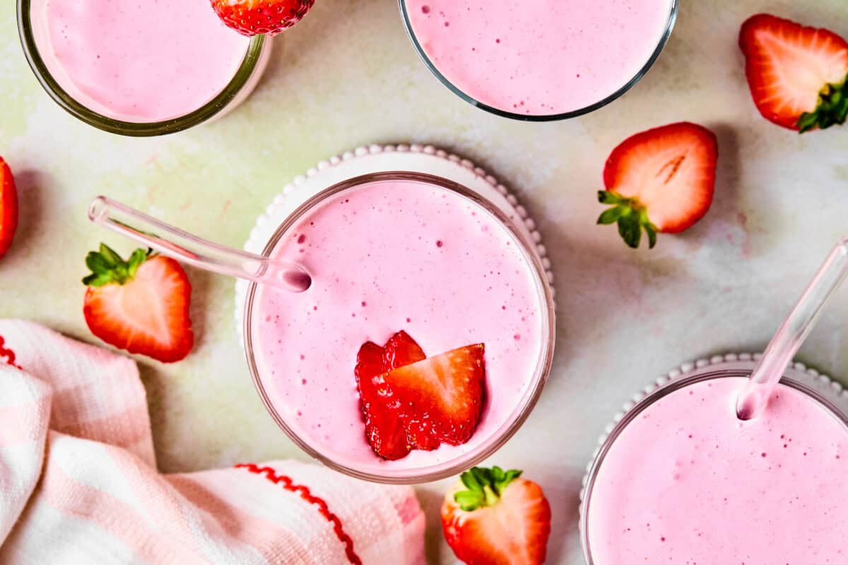 Top-down view of glasses filled with pink strawberry protein shake, each with a clear straw and strawberry slices on top. Fresh strawberries and a white cloth with red trim are scattered on a light surface.