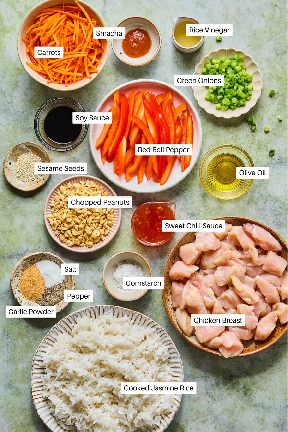 Top-down view of labeled ingredients for sweet chili chicken stir-fry, including sliced carrots, rice vinegar, sriracha, soy sauce, red bell pepper, green onions, sesame seeds, peanuts, sweet chili sauce, cornstarch, chicken, oil, salt, pepper, garlic powder, and rice.