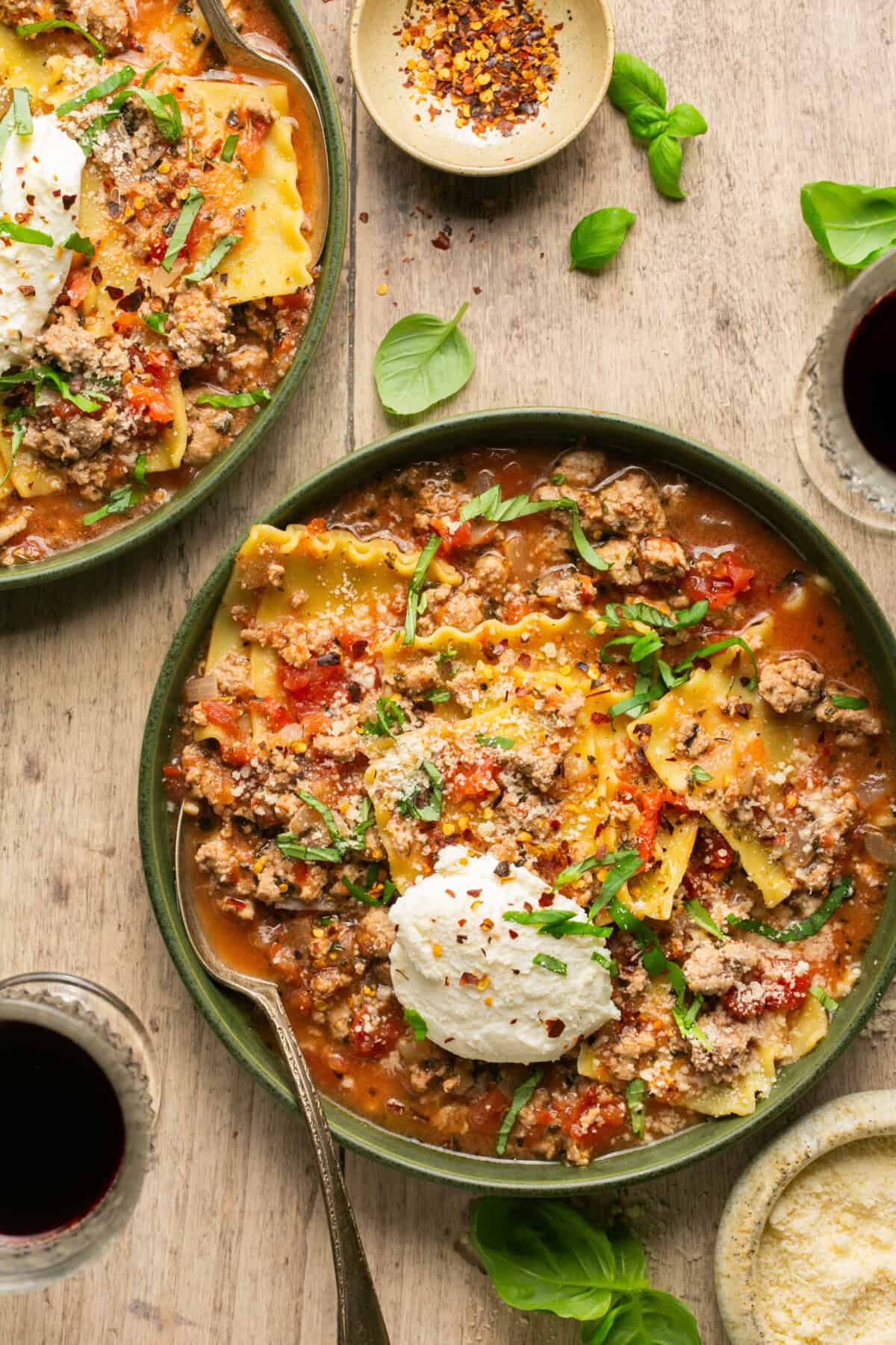 A bowl of lasagna soup with ground meat, tomatoes, pasta sheets, fresh basil, and ricotta cheese—a perfect addition to your winter comfort foods—served alongside red pepper flakes, grated cheese, and glasses of red wine on a wooden table.