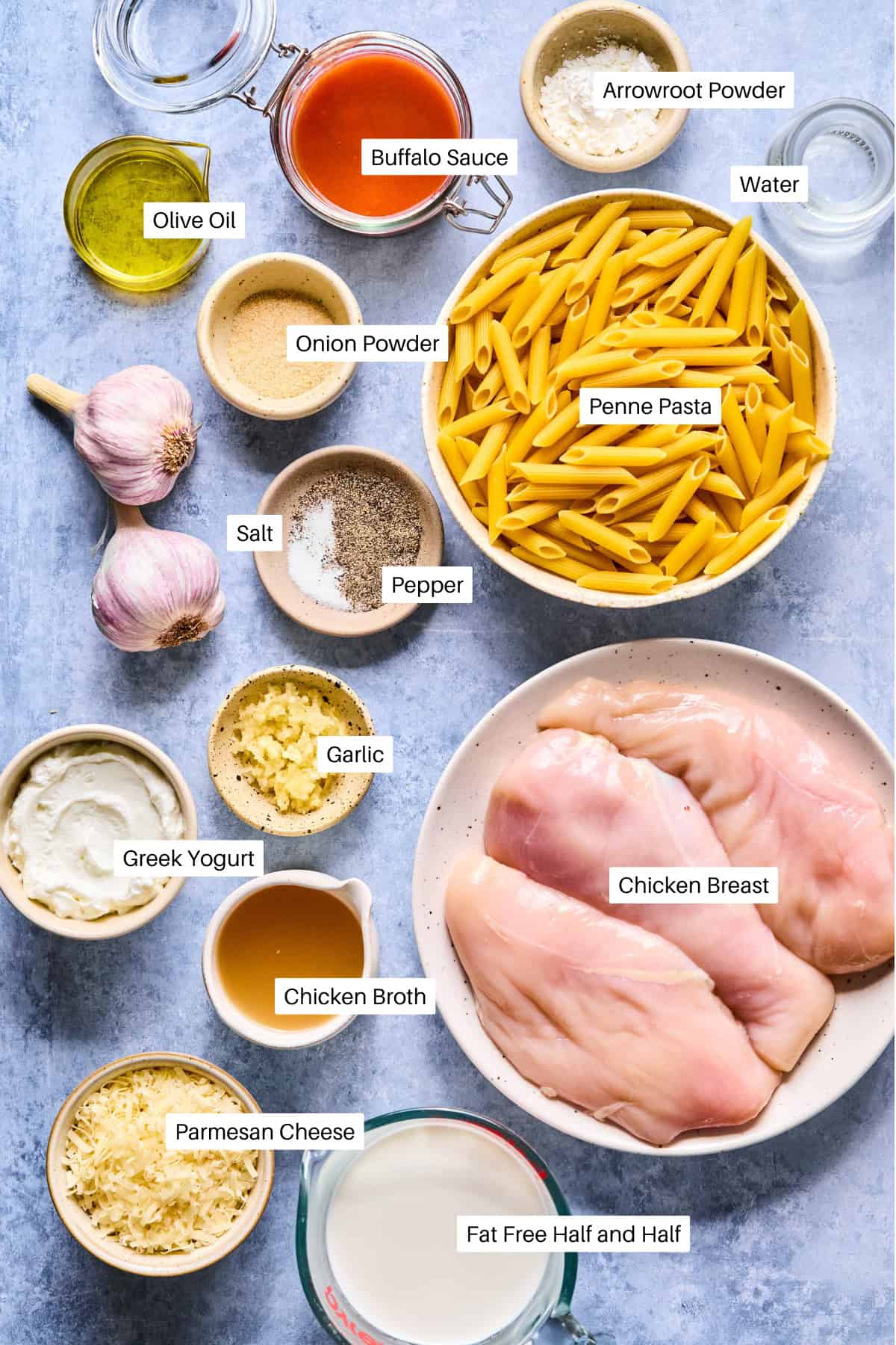 Labeled ingredients for Buffalo Chicken Pasta are laid out on a blue surface, including penne pasta, chicken breasts, Greek yogurt, parmesan cheese, garlic, olive oil, buffalo sauce, half and half, chicken broth, arrowroot powder, and seasonings.