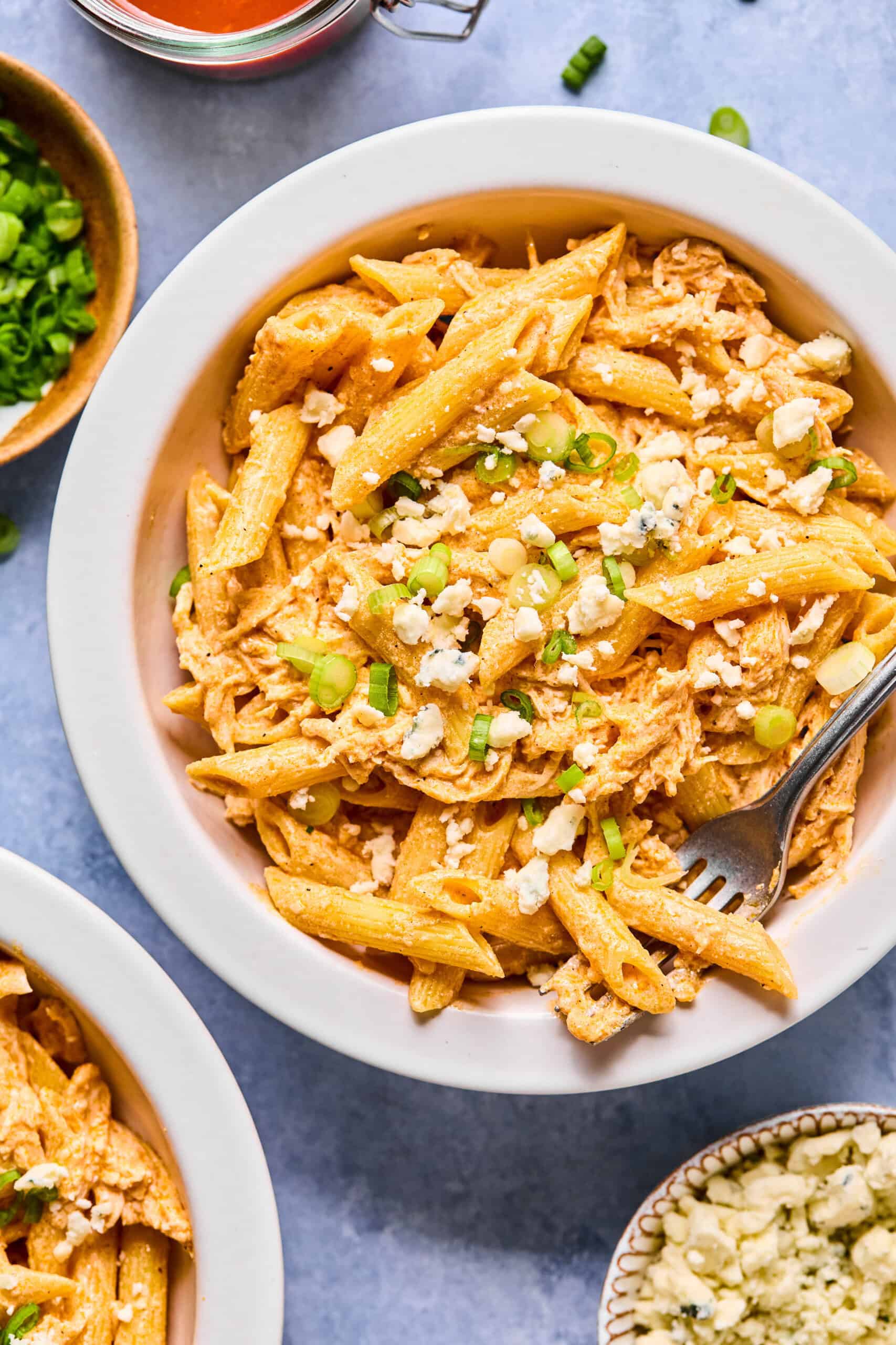 A bowl of creamy Buffalo Chicken Pasta topped with crumbled cheese and chopped green onions, with a fork resting in the bowl. Small bowls of sauce, green onions, and cheese are nearby on a blue surface.
