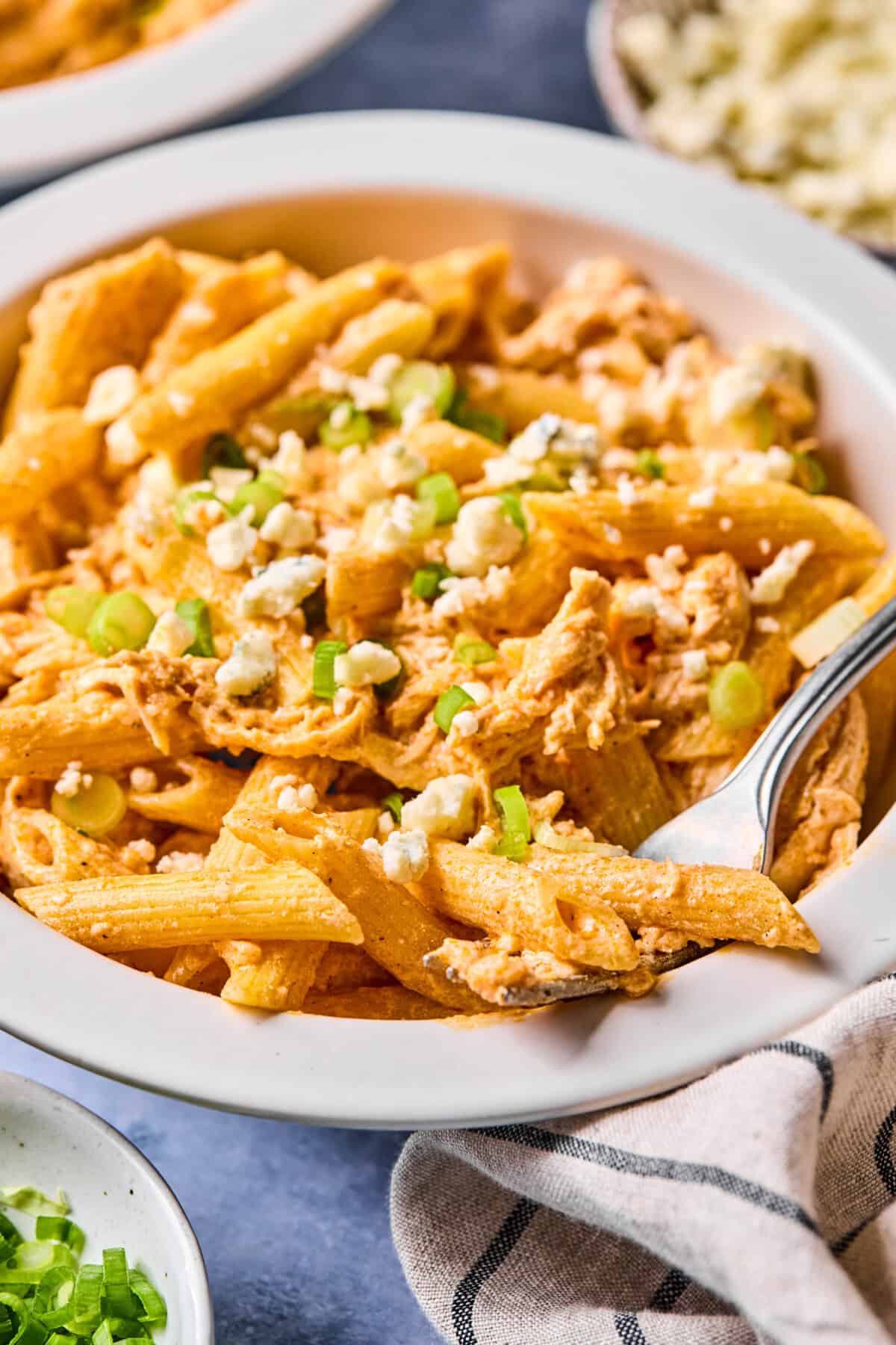 A bowl of creamy Buffalo Chicken Pasta mixed with shredded chicken, topped with chopped green onions and crumbled cheese, with a fork resting in the bowl. A cloth napkin and small bowls are nearby on the table.