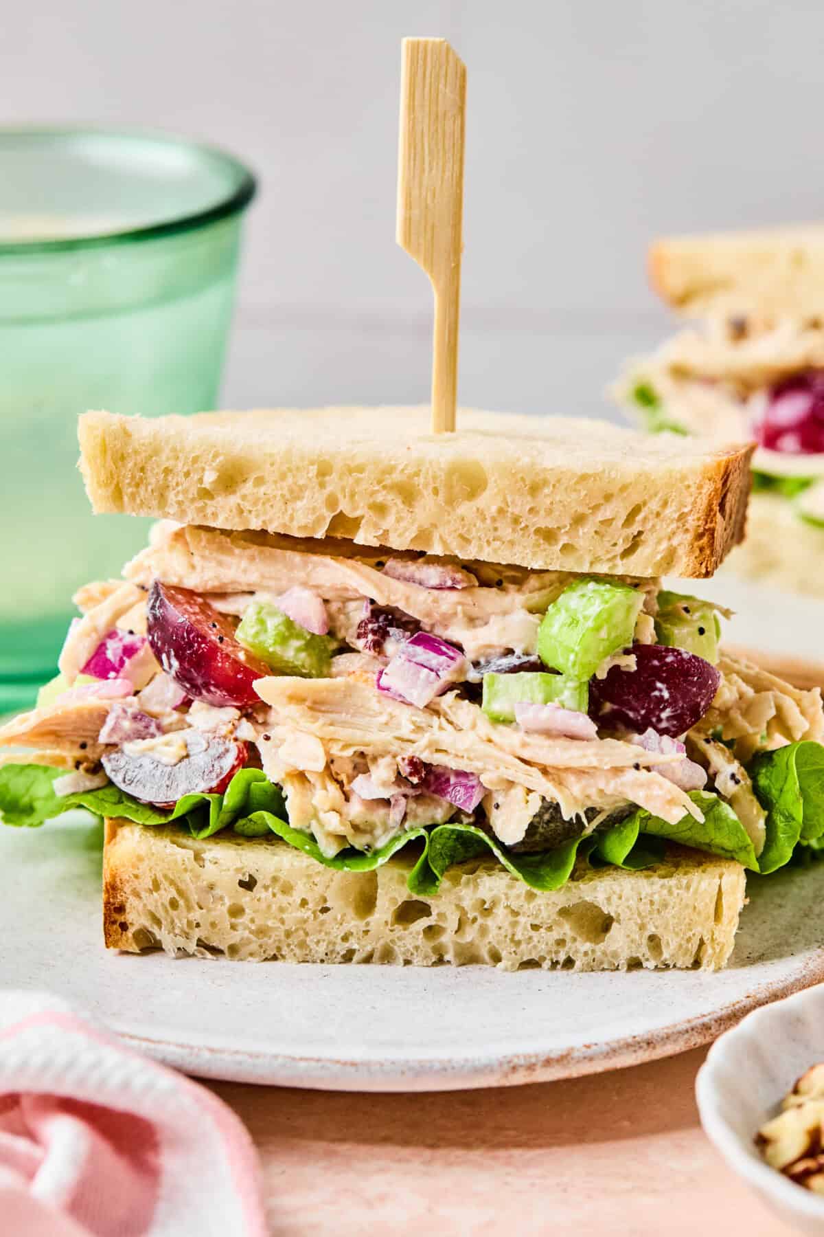 A California Chicken Salad sandwich with lettuce, red onions, celery, and red grapes on sliced bread, held together by a wooden skewer, sits on a white plate. A light green glass and another sandwich are blurred in the background.