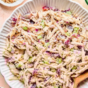 A large white bowl filled with creamy California Chicken Salad featuring shredded chicken, celery, red onions, grapes, and cranberries, with a wooden spoon resting in the bowl. Small dishes of pecans and drinks are nearby.