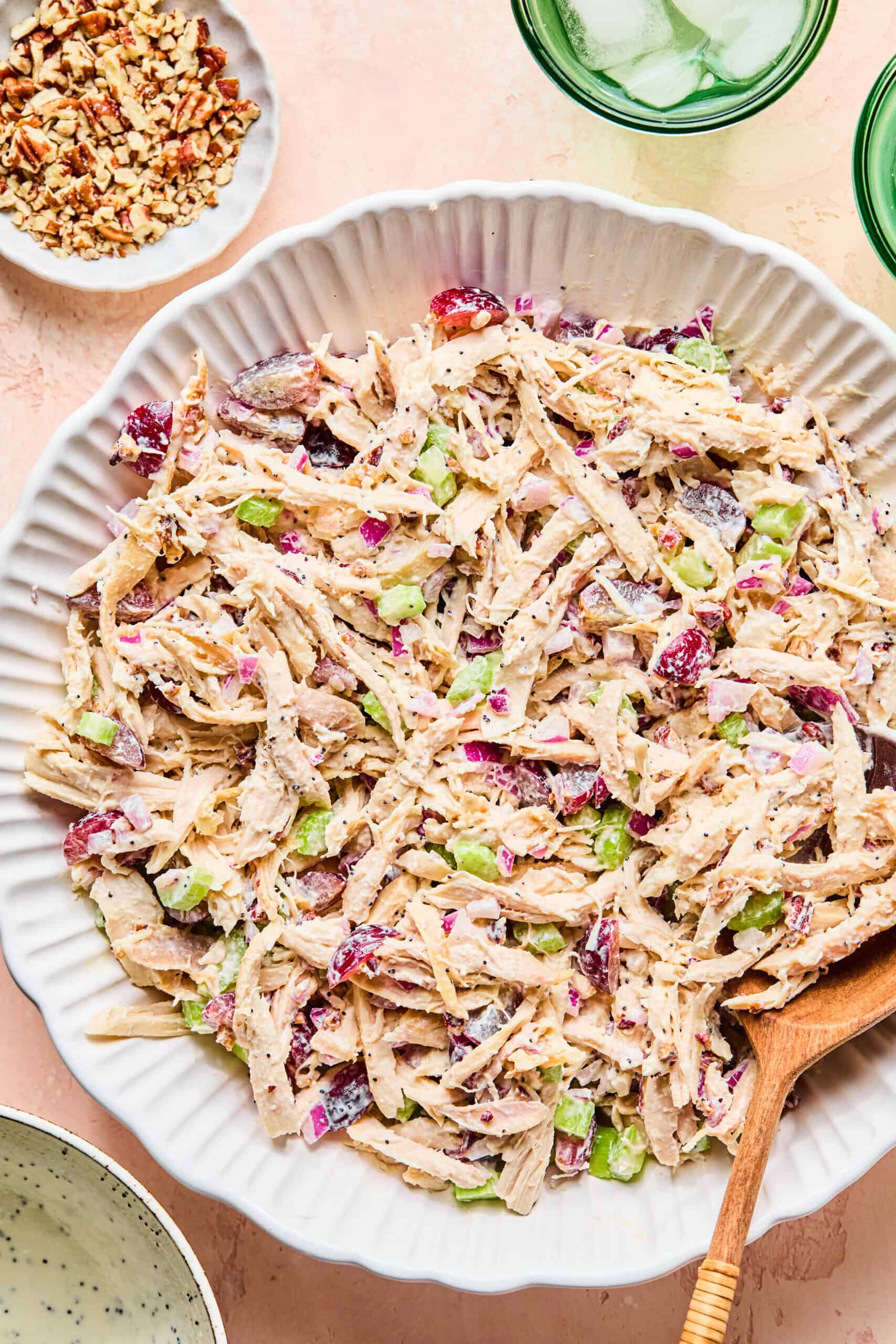 A large white bowl filled with creamy California Chicken Salad featuring shredded chicken, celery, red onions, grapes, and cranberries, with a wooden spoon resting in the bowl. Small dishes of pecans and drinks are nearby.