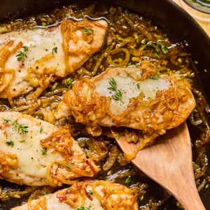 A skillet with French Onion Chicken: chicken breasts topped with melted cheese, caramelized onions, and fresh herbs. A wooden spatula lifts one piece; the dish is golden and savory with visible onions and herbs.