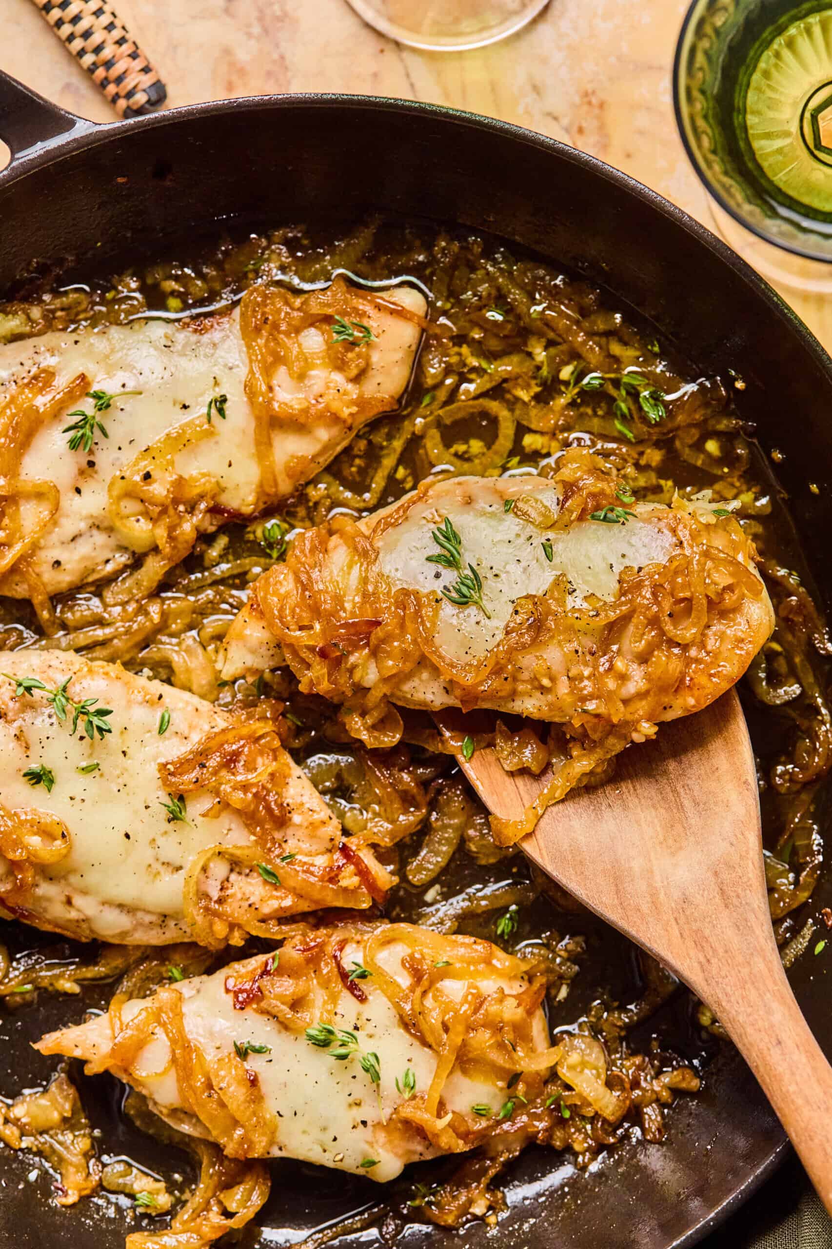 A skillet with French Onion Chicken: chicken breasts topped with melted cheese, caramelized onions, and fresh herbs. A wooden spatula lifts one piece; the dish is golden and savory with visible onions and herbs.