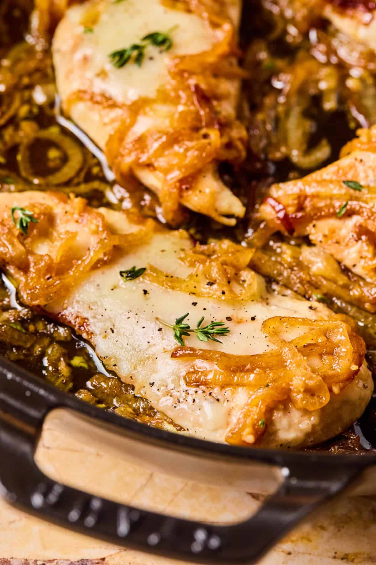 A close-up of French Onion Chicken in a skillet, featuring cooked chicken breasts topped with melted cheese, caramelized onions, and fresh thyme sprigs. The dish sits in a savory-looking sauce.