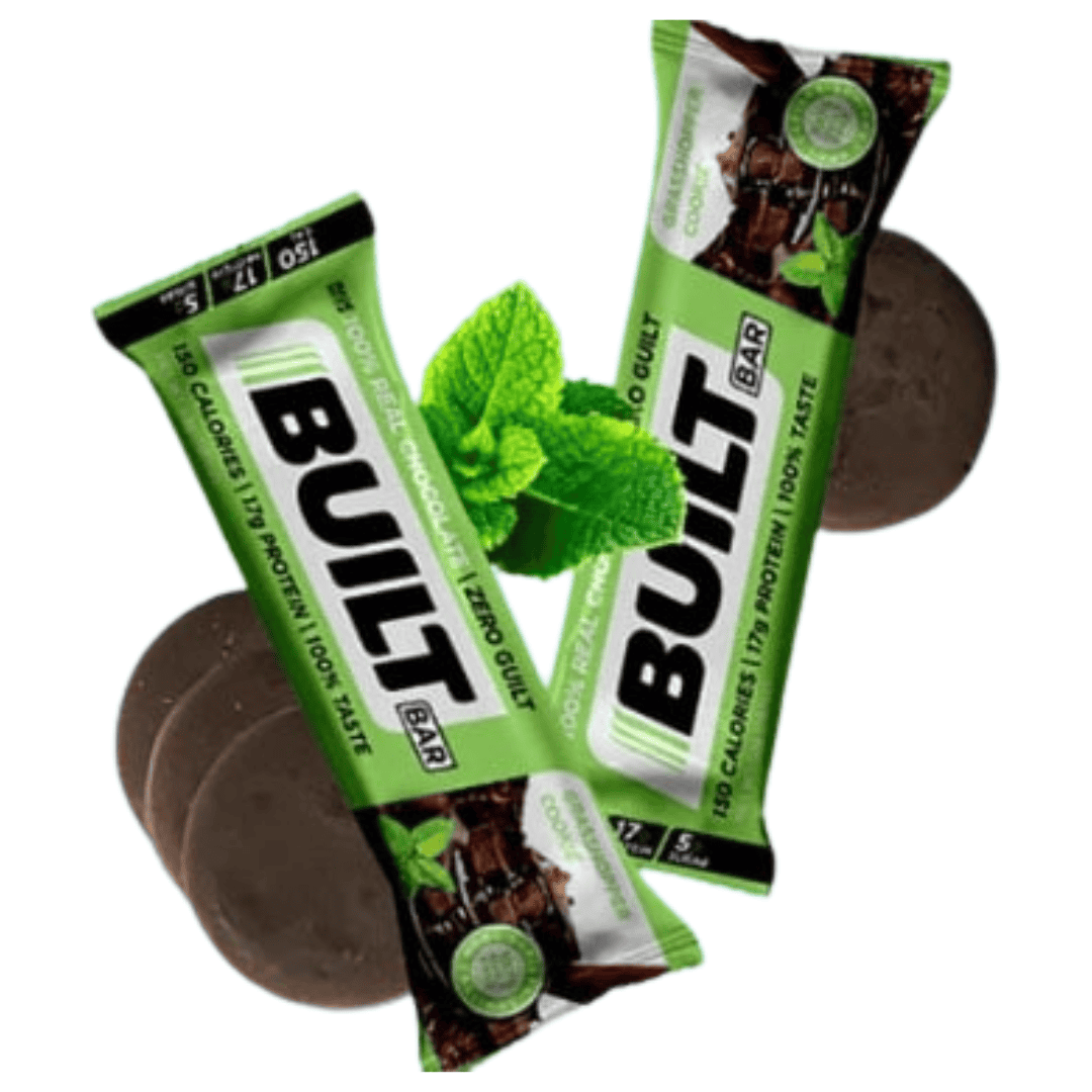 Two Built Bar protein bars in green mint chocolate chip packaging with fresh mint leaves and chocolate-coated protein bars in the background.