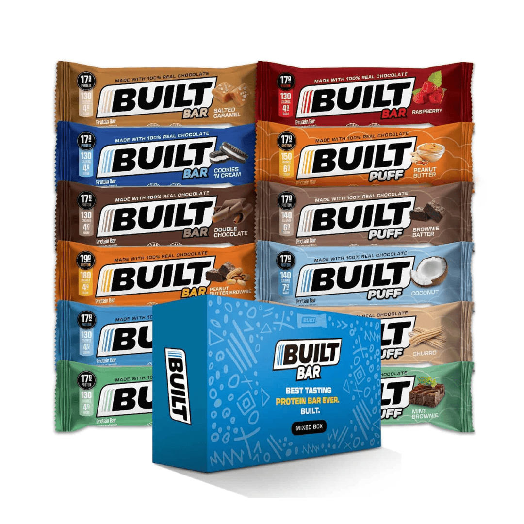 Nine different flavors of Built Bar protein bars are arranged standing upright behind a blue Built Bar Mixed Box, which is labeled Best Tasting Protein Bar Ever. Each bar has colorful packaging.