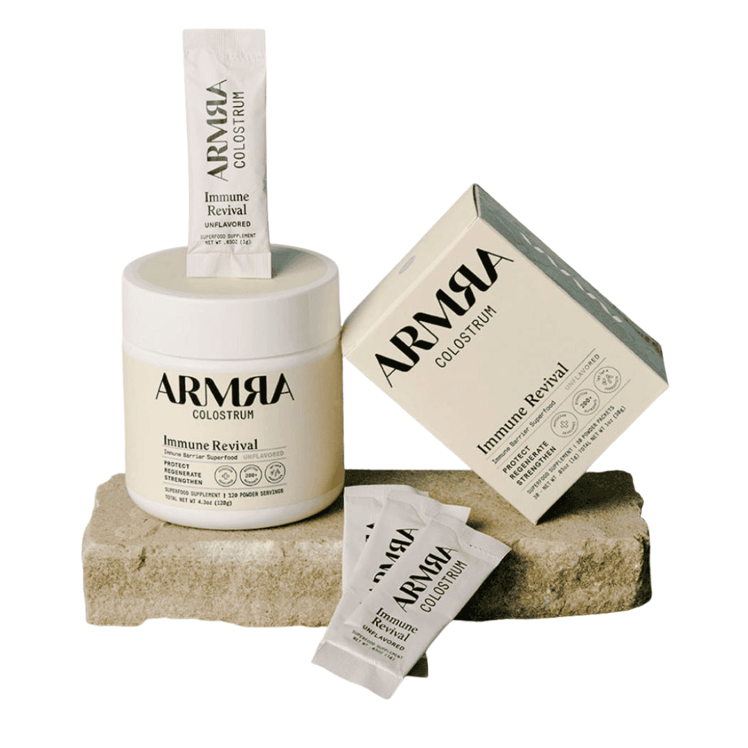 A container and box of ARMRA Colostrum Immune Revival supplement, with several single-serve packets, displayed on a textured stone against a white background.