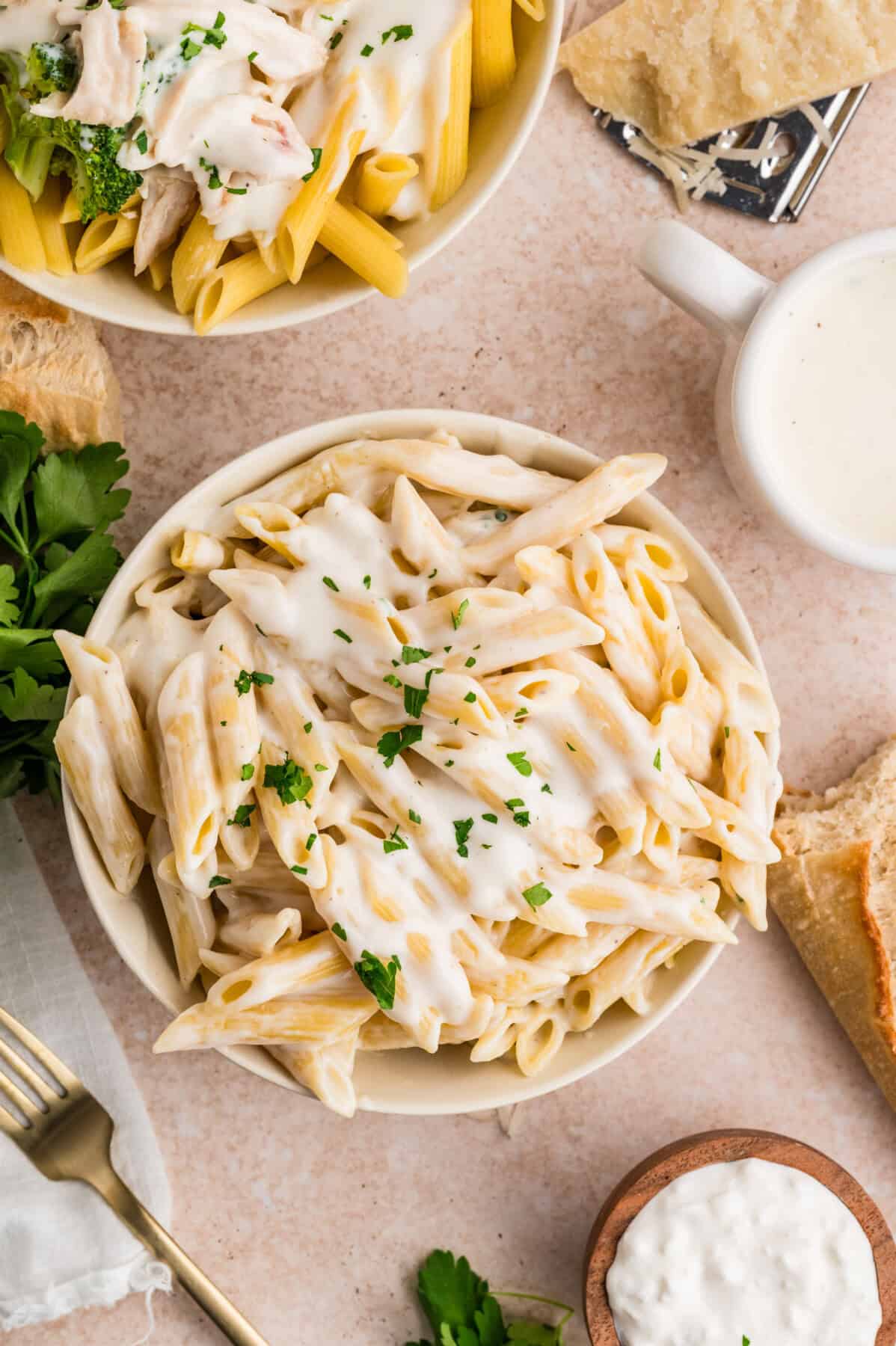 A bowl of penne pasta topped with creamy cottage cheese Alfredo sauce and garnished with chopped parsley, surrounded by bread, a fork, fresh herbs, and another bowl of pasta on a light surface.