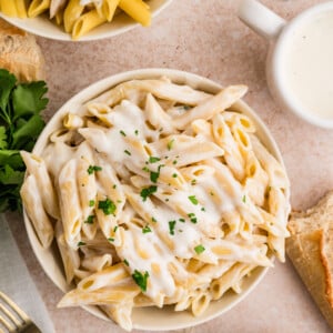 A bowl of penne pasta topped with creamy cottage cheese Alfredo sauce and garnished with chopped parsley, surrounded by bread, a fork, fresh herbs, and another bowl of pasta on a light surface.