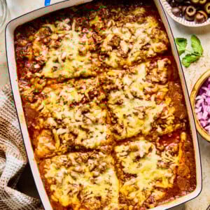 A baked casserole dish of cheesy lasagna cut into six large squares, topped with melted cheese and meat sauce—this twist on lazy enchiladas is surrounded by bowls of chopped herbs, sliced black olives, and diced red onions.