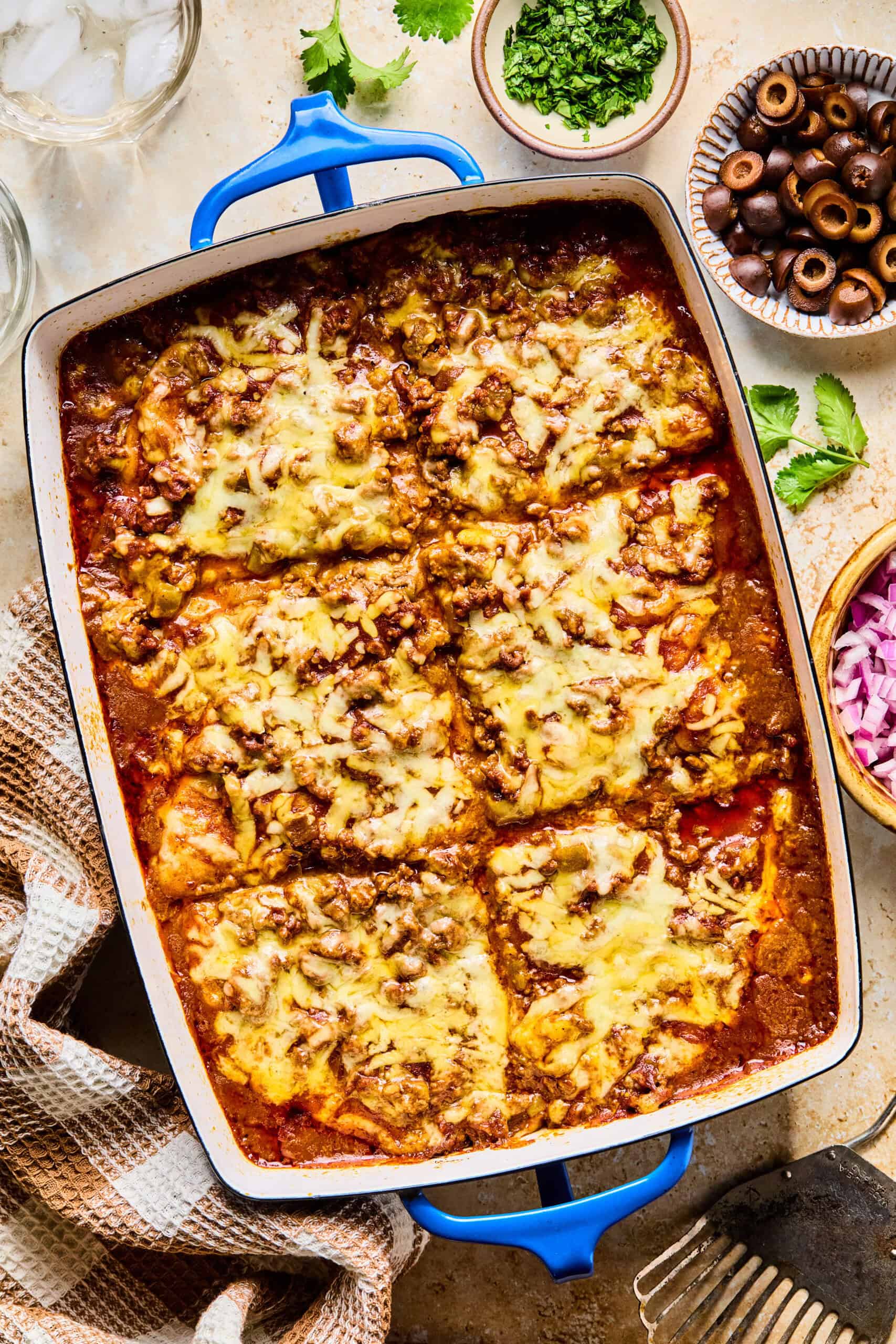 A baked casserole dish of cheesy lasagna cut into six large squares, topped with melted cheese and meat sauce—this twist on lazy enchiladas is surrounded by bowls of chopped herbs, sliced black olives, and diced red onions.