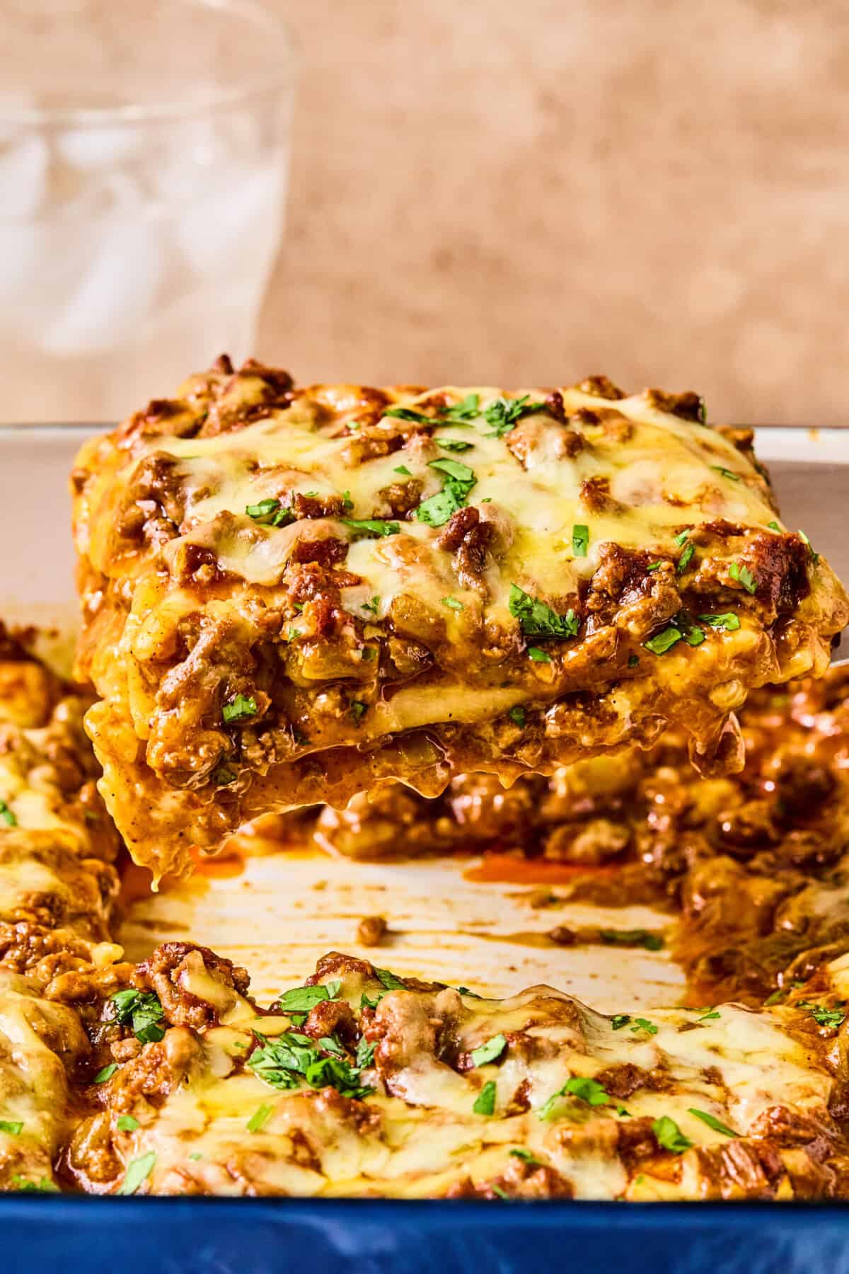 A close-up of a cheesy lasagna slice being lifted from a baking dish, with visible layers of noodles, meat sauce, and melted cheese—perfect for fans of lazy enchiladas. A glass of ice water is in the blurred background.