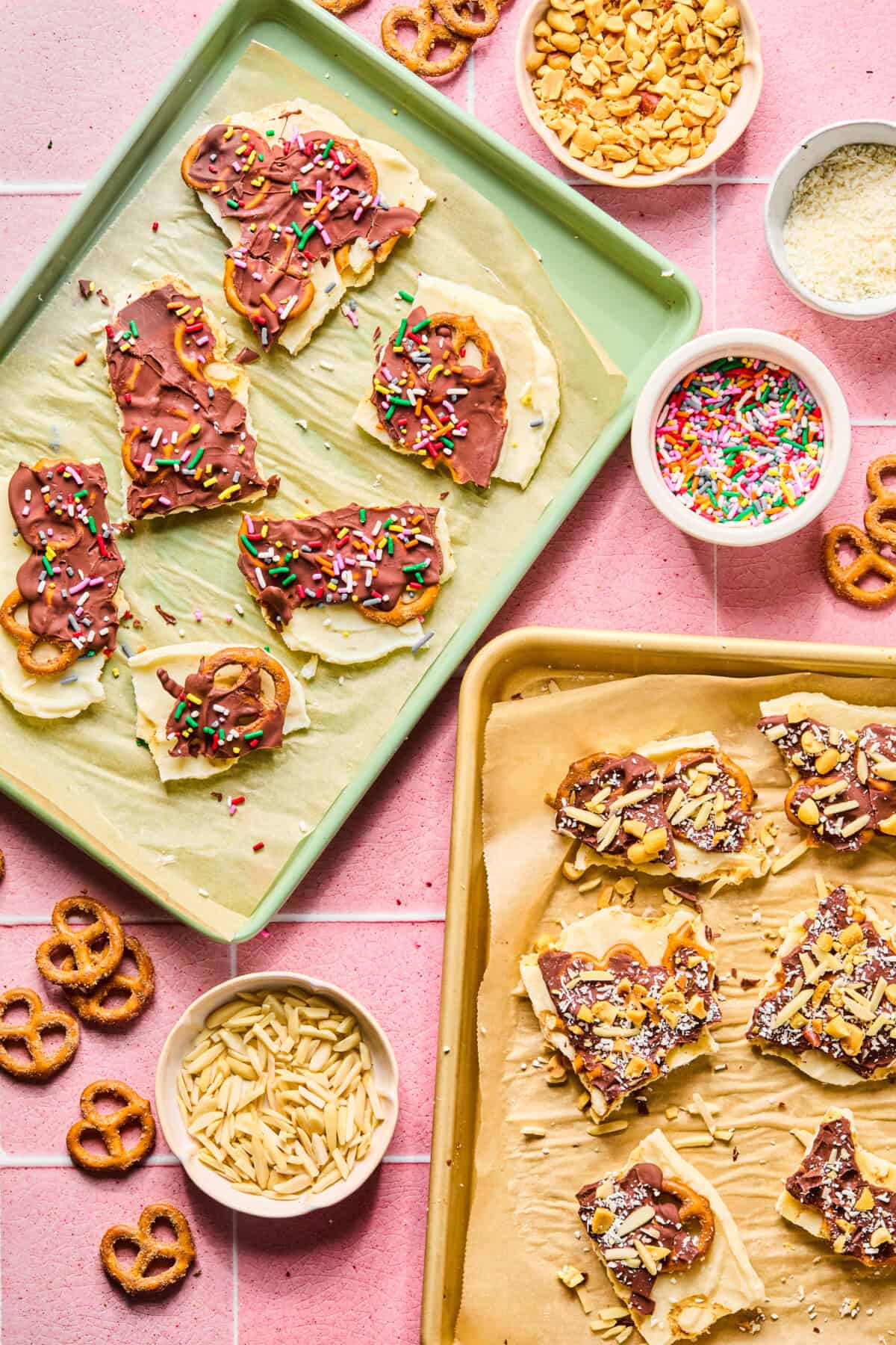 Two trays of chocolate-dipped pretzel bark topped with colorful sprinkles and nuts sit on a pink tiled surface, surrounded by bowls of sprinkles, slivered almonds, chopped nuts, pretzels, and protein frozen yogurt bark pieces.