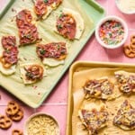 Two trays of chocolate-dipped pretzel bark topped with colorful sprinkles and nuts sit on a pink tiled surface, surrounded by bowls of sprinkles, slivered almonds, chopped nuts, pretzels, and protein frozen yogurt bark pieces.