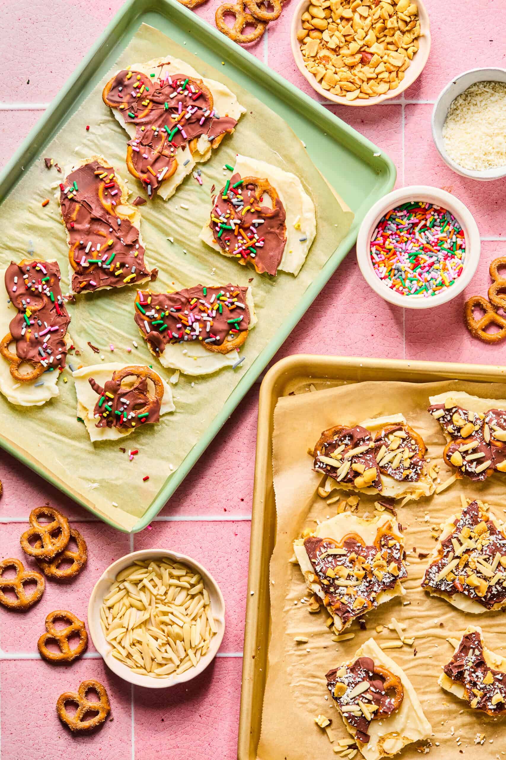 Two trays of chocolate-dipped pretzel bark topped with colorful sprinkles and nuts sit on a pink tiled surface, surrounded by bowls of sprinkles, slivered almonds, chopped nuts, pretzels, and protein frozen yogurt bark pieces.