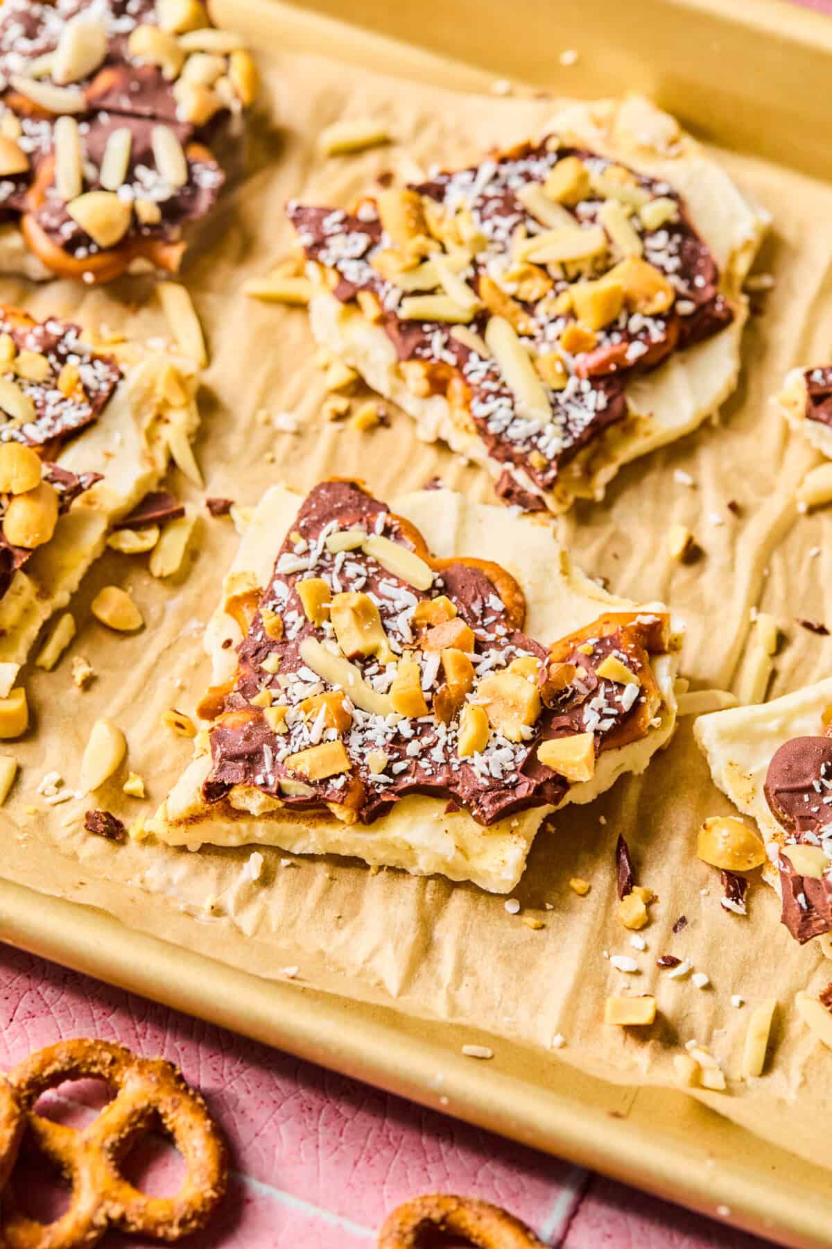 Squares of dessert bark with a cracker base, topped with chocolate, chopped nuts, and shredded coconut, are arranged on a parchment-lined baking sheet. Inspired by protein frozen yogurt bark, pretzel pieces add a crunchy twist at the edge of the image.