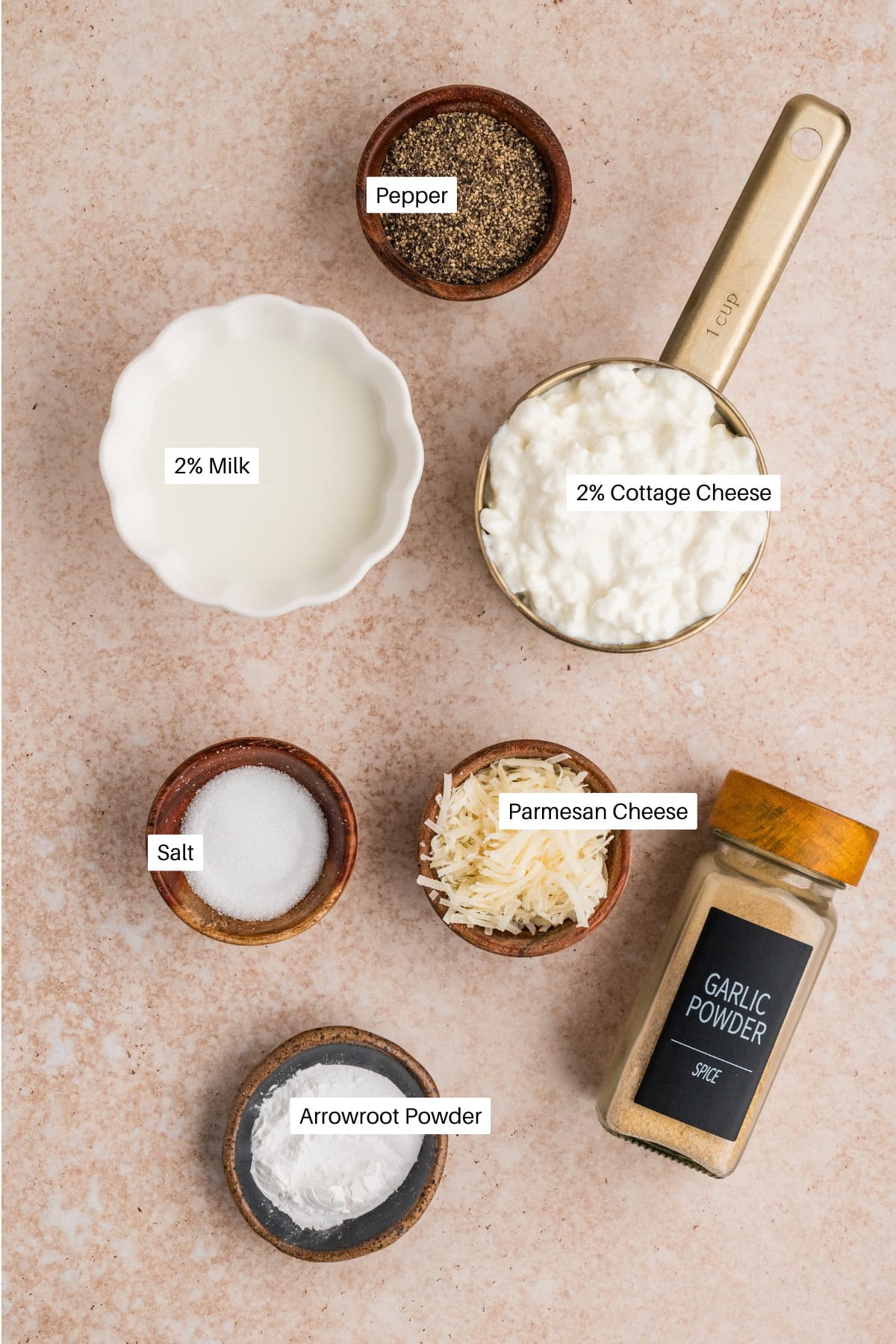 Ingredients arranged on a beige surface: a ramekin of 2% milk, a gold cup of 2% cottage cheese for creamy cottage cheese alfredo, bowls of pepper, salt, shredded Parmesan cheese, arrowroot powder, and a jar of garlic powder.