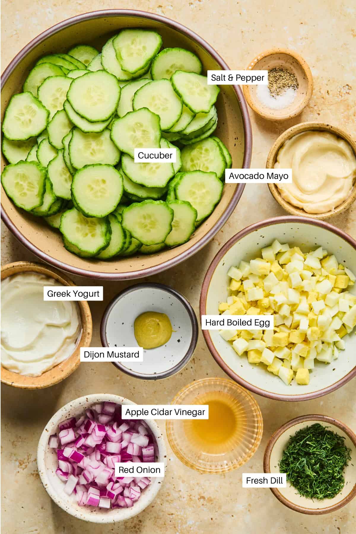 A top-down view of bowls holding ingredients for a cucumber and dill salad: sliced cucumber, avocado mayo, Greek yogurt, diced hard-boiled egg, Dijon mustard, apple cider vinegar, chopped red onion, fresh dill, salt, and pepper. Labels identify each item.