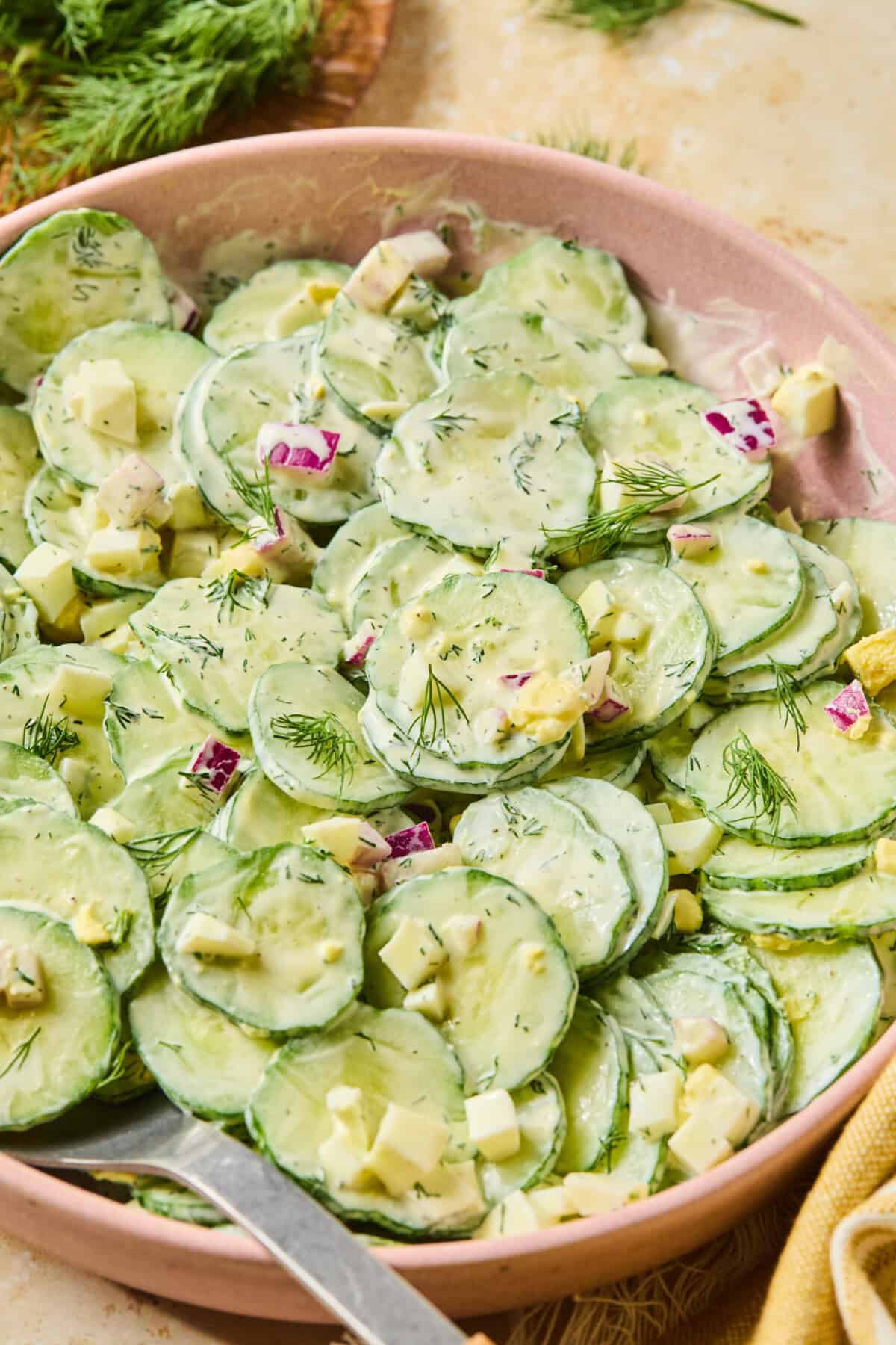 A bowl of creamy cucumber and dill salad with thinly sliced cucumbers, diced cheese, red onions, and fresh dill, served with a large spoon.