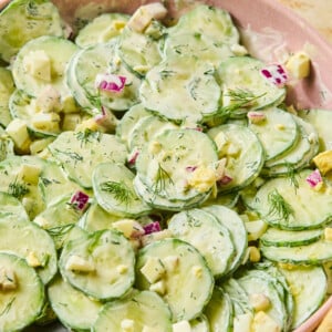 A bowl of creamy cucumber and dill salad with thinly sliced cucumbers, diced cheese, red onions, and fresh dill, served with a large spoon.