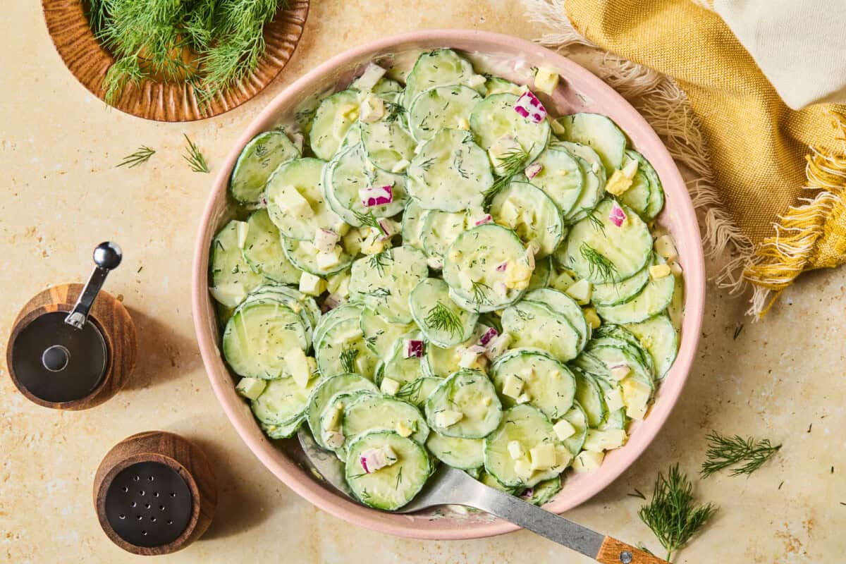 A bowl of creamy cucumber and dill salad with red onion and diced cheese, served on a beige surface with a yellow cloth, pepper and salt shakers, and a small plate of fresh dill nearby.