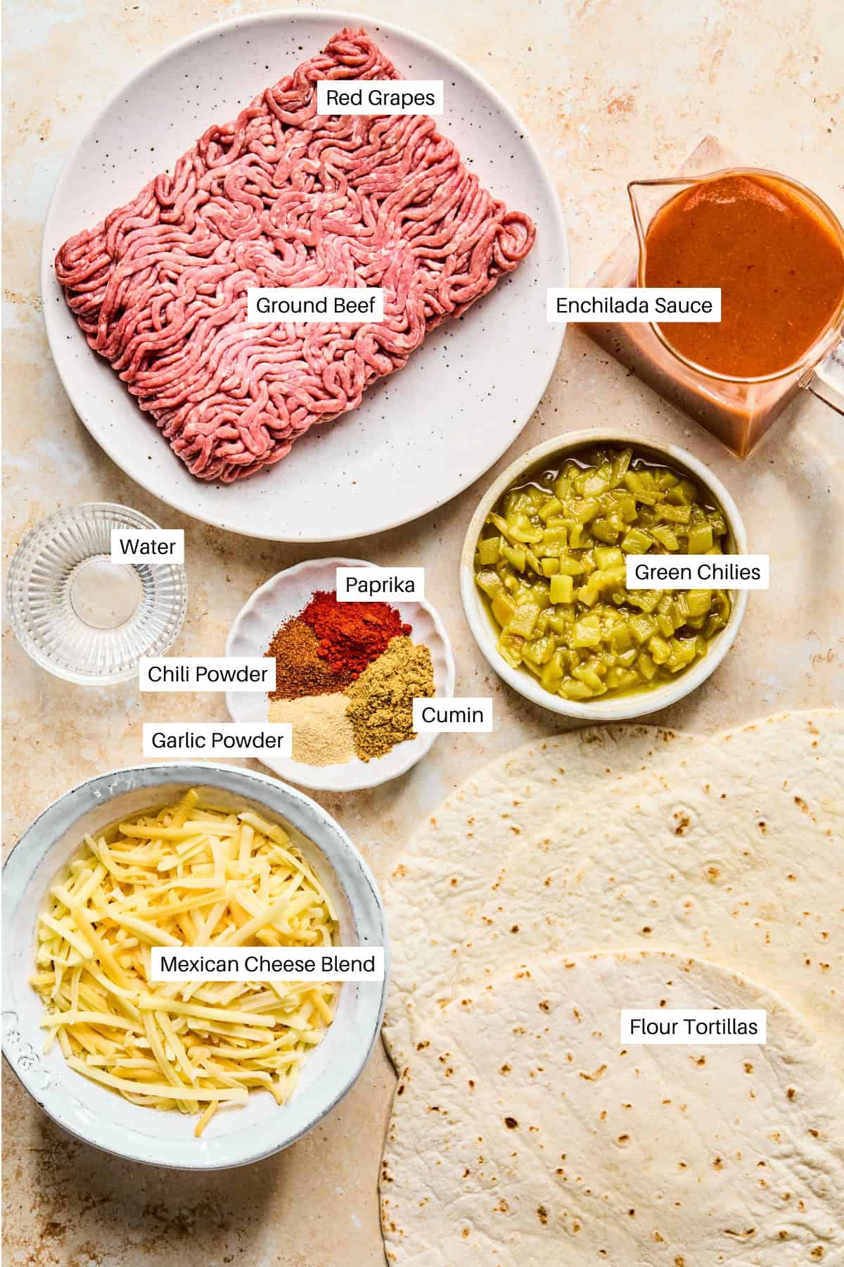 A flat lay of labeled ingredients for lazy enchiladas: ground beef, enchilada sauce, green chilies, spices, Mexican cheese blend, and flour tortillas. One label, Red Grapes, is mistakenly placed above the beef.