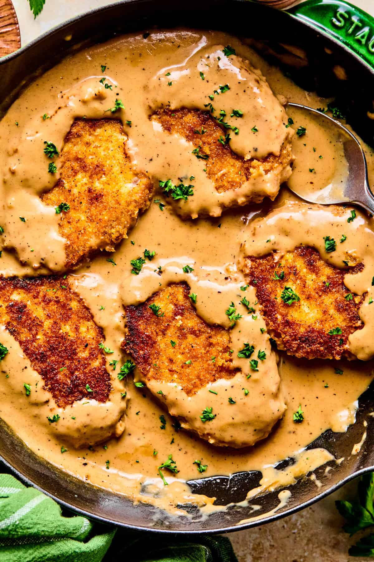 Four golden-brown, breaded pork chops and gravy simmer in a skillet, partially covered with creamy brown sauce and sprinkled with chopped parsley. A large spoon rests beside the cutlets in the rich, savory mixture.
