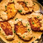 Four golden-brown, breaded pork chops and gravy simmer in a skillet, partially covered with creamy brown sauce and sprinkled with chopped parsley. A large spoon rests beside the cutlets in the rich, savory mixture.