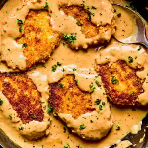 Four golden-brown, breaded pork chops and gravy simmer in a skillet, partially covered with creamy brown sauce and sprinkled with chopped parsley. A large spoon rests beside the cutlets in the rich, savory mixture.